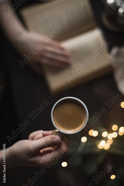 Fototapeta Cup of espresso in the girl's hand and a book, dark photo, selective focus, cozy evening photo, top view