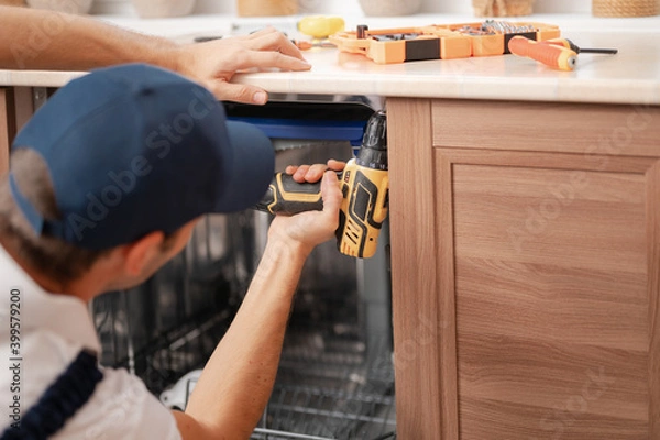 Fototapeta A man or a service worker in special clothes using a screwdriver attaches to the countertop or removes the dishwasher, close-up
