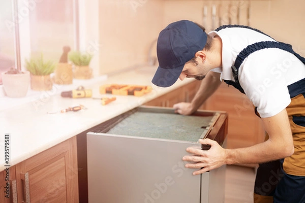 Obraz A man or service worker in special clothing installs, disassembles or performs maintenance of the built-in dishwasher