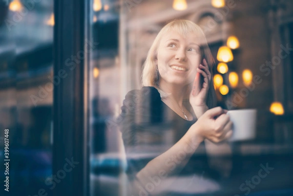 Fototapeta Beautiful young blonde woman sitting in a cafe talking on a mobile phone by the window with a reflection of the city street