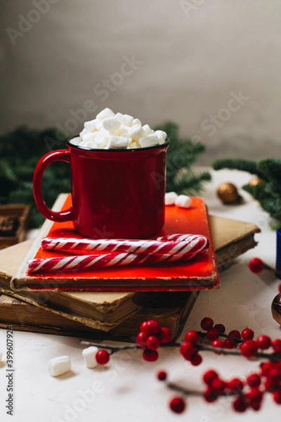 Obraz A red mug with cocoa and marshmallows stands on a stack of old books. Nearby are spruce branches, New Year's decorations, a branch of ikles and stamps for letters. Christmas mood and a hot drink.