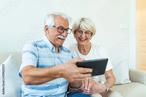 Fototapeta Couple of seniors smiling and looking at the same tablet hugged on the sofa - indoor, at home concept - caucasians mature and retired man and woman using technology