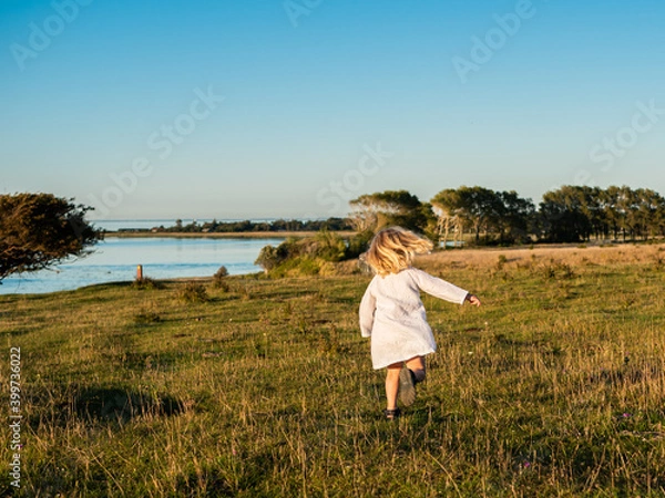 Fototapeta Blond girl 4 years old in white summer dress running and dancing on a field in sunlight and blue sky above