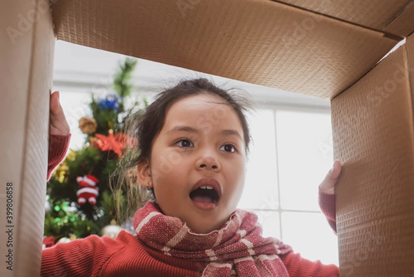 Fototapeta merry christmas and happy holidays. cheerful cute child girl opening a christmas present. little kid having fun near christmas tree indoors. view from inside of the box.