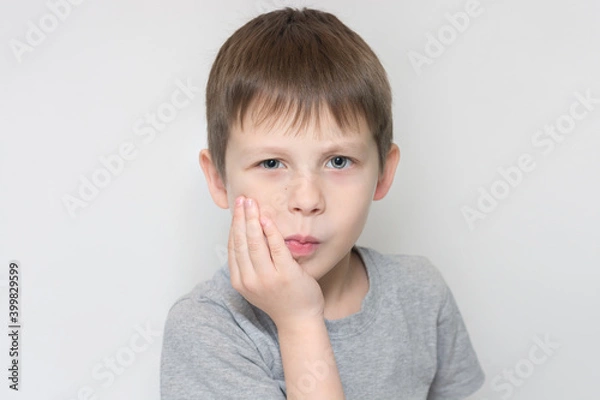 Fototapeta the child holds his cheek with his hand, indicating that a tooth hurts. The boy looks at the camera, horizontal photo, portrait. Unhappy preschooler with toothache.