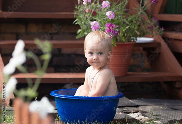 Fototapeta a little girl is sitting in a blue basin of water on the street near the house, bathing and something is very surprised