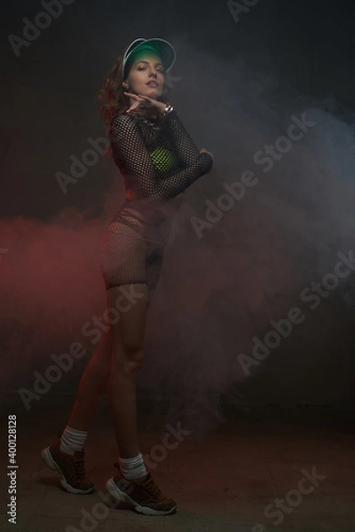 Fototapeta Portrait of a charming and stylish girl with slim figure weared with green cap and dressed in reticulated clothing poses in dark smokey background.