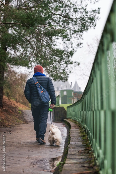 Obraz A man in a down jacket and his dog walk along a path with a green railing towards a castle.