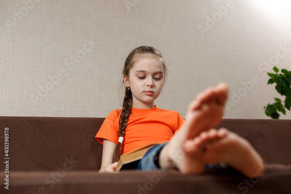 Fototapeta A little caucasian girl with bare feet reading book while sitting on a sofa at home. selective focus