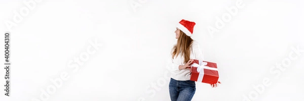 Fototapeta Young beautiful woman in red santa hat and white sweater holding gift box and looking to the side isolated on white background. Banner size, empty space