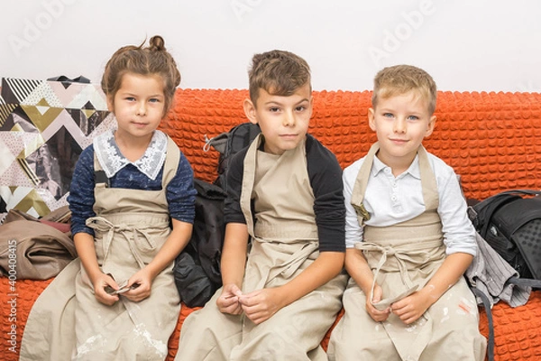 Fototapeta Three children sit in aprons on a red sofa and look at the camera