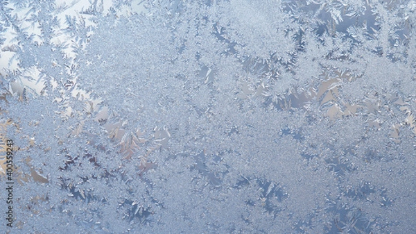 Fototapeta Ice and frost on the window glass in winter. A pattern of frozen water crystals, similar to the leaves of fantastic plants or jewels. Light blue backdrop. Abstract natural background or wallpaper