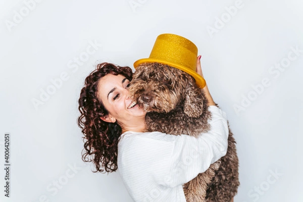 Fototapeta A beautiful woman having fun with her Spanish water dog in a party at home. She is putting a golden top hat on the dog's head. New year's eve with dogs at home concept