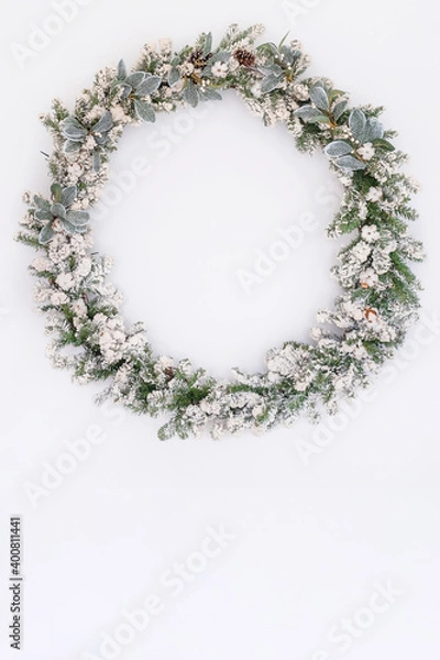 Fototapeta Snowy big christmas wreath hanging on white wall indoors, festive decoration. Creative wreath with cones, cotton flowers, mistletoe, isolated in white. Handmade holiday decor. Copy space. Mockup