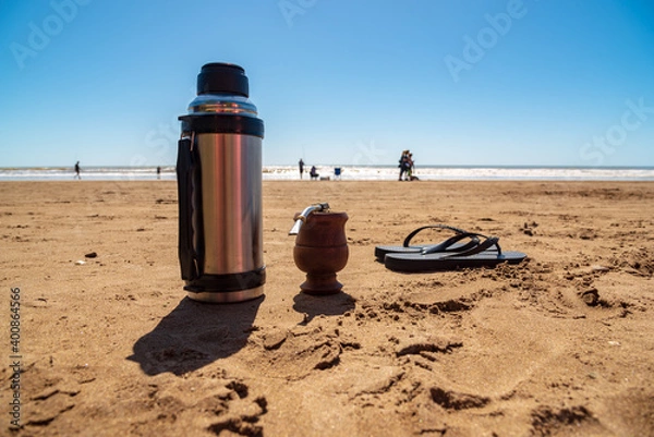 Fototapeta A thermos with a mate and a pair of flip flops resting on the sand on the beach with people walking in the background