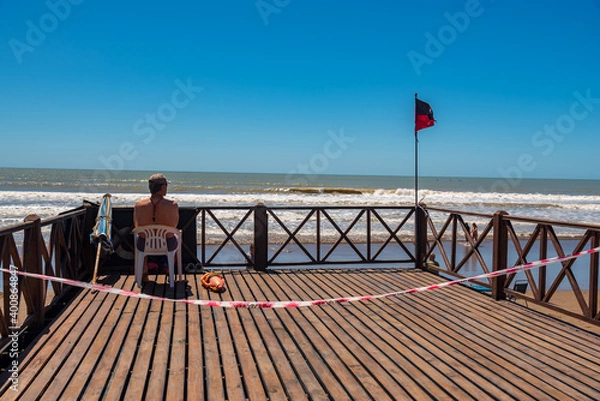 Fototapeta Mar del Tuyú, Buenos Aires, Argentina; December 06, 2020: Lifeguard sitting on a deck next to the tidal flag. A do not cross link because social distancing during COVID-19