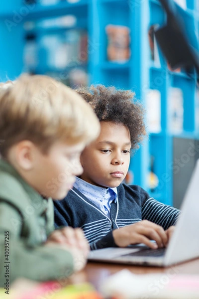 Obraz Side view of two boys working on laptop computer sitting at desk in school library. Focus on curly-haired mixed race boy typing on keyboard