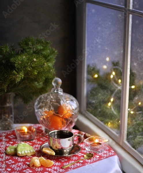 Fototapeta A cup of coffee, tangerines, cookies and fir branches on the table near the snowy window. Lights and candles. Snow outside the window.