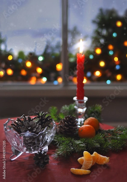 Fototapeta A red candle, fir branches, tangerines and cones on the table near a snowy window. Lights outside the window.