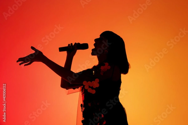 Fototapeta Talented. Silhouette of young female singer isolated on orange gradient studio background in neon light. Beautiful shadow in action, performing. Concept of human emotions, expression, ad, music, art.