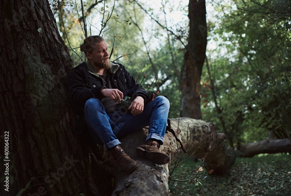 Fototapeta Portrait of a smoking young bearded man adventurer relaxing in the autumn forest and holding a cigarette