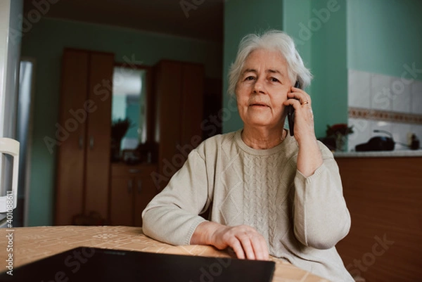 Fototapeta Portrait of a senior grandmother talking on the phone with her family while sitting at a table in the house. Communication during isolation of COVID-19 coronavirus