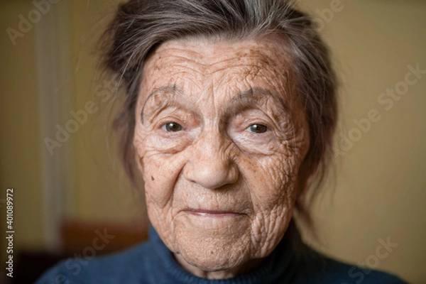 Fototapeta 90 year old cute elderly woman with gray hair and wrinkles face, wearing sweater, portrait large, smiling and looking joyfully, background of room. Theme long-liver and aging, old people in good mood