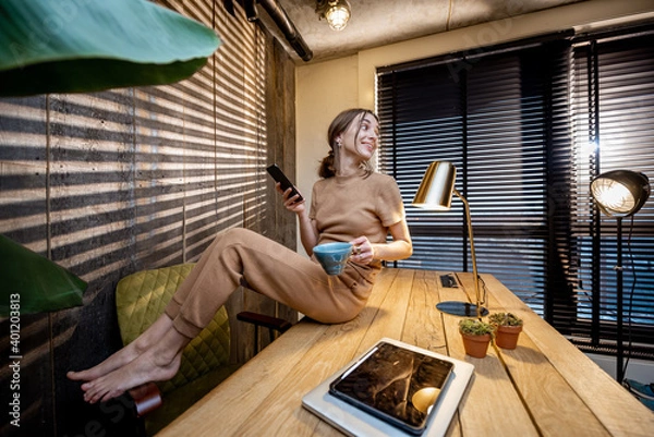Fototapeta Relaxed woman in comfortable domestic suit with a smart phone and coffee cup sitting on the table at the beautiful home office