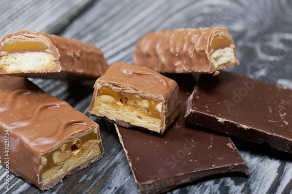 Obraz Chopped up chocolate bar. Filled with caramel and nuts. Nearby are pieces of chocolate. On black pine boards.