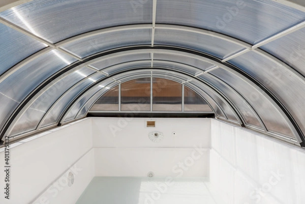 Fototapeta An empty fiberglass swimming pool with a sliding polycarbonate cover on a sunny spring day. A pool is white-colored, while the pavilion is half-transparent with blue toning.