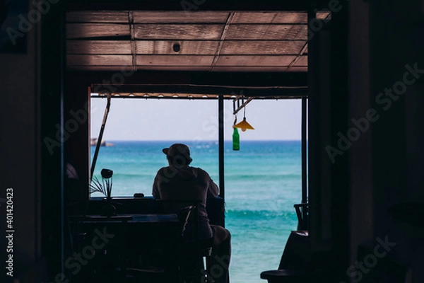Fototapeta A silhouette of a man, sitting in a vintage style cafe on a beach in front of blue ocean panorama, enjoying vacation moment. Travel relax concept background.
