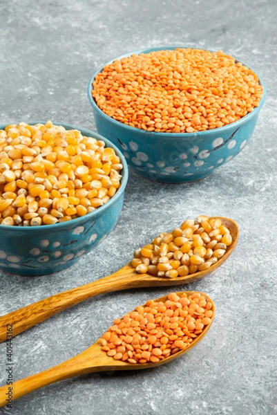 Fototapeta Two bowls of raw corn kernels and red lentil on marble surface