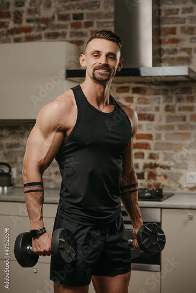 Obraz A happy muscular man with a beard is posing with dumbbells in his apartment. An athletic guy with tattoos on his arms is warming up during a workout at home. An athlete is training his biceps.