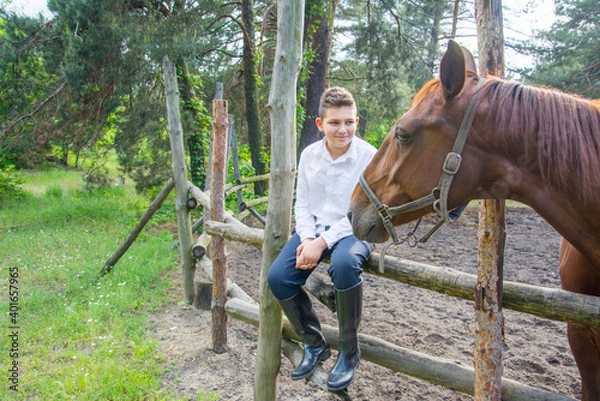 Fototapeta In summer, on a bright sunny day, a boy is sitting next to a horse on a ranch.