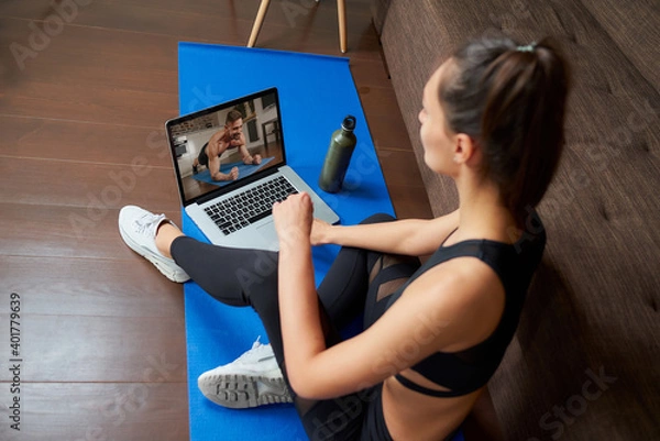 Obraz A laptop screen view over a woman's shoulder. A sporty girl in a tight suit is watching a workout video on laptop in her apartment. Woman is listening to a coach during a virtual fitness class at home