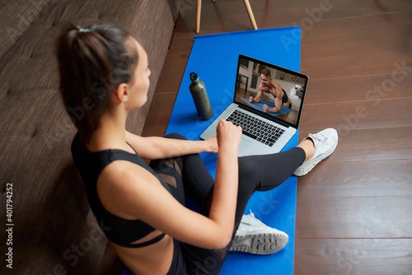 Obraz A laptop screen view over a woman's shoulder. A sporty girl in a tight suit is watching a workout video on laptop in her apartment. Woman is listening to a coach during a virtual fitness class at home