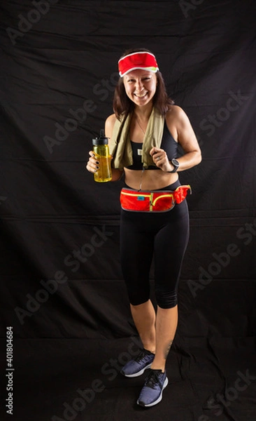 Fototapeta Beautiful girl in jogging red uniform, drinks water after training in the studio on a black background