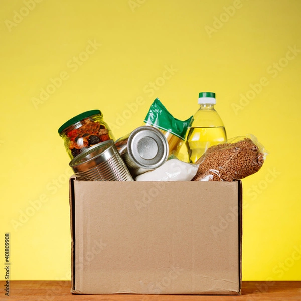 Fototapeta Food Donation concept. Donation Box With food For Donation On Yellow. Assistance To The Elderly In The Context of The Coronavirus Pandemic. square image
