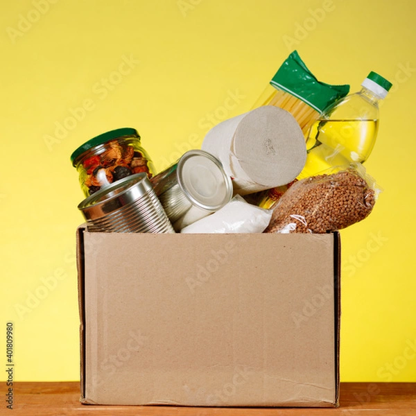 Fototapeta Food Donation concept. Donation Box With food For Donation On Yellow. Assistance To The Elderly In The Context of The Coronavirus Pandemic. square image