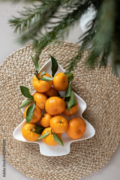 Fototapeta Small ripe tangerines with leaves, on plate in shape of Christmas tree on wicker serving napkin. Concept winter vitamin food. Moody still life festive composition. For greeting card, book, stories