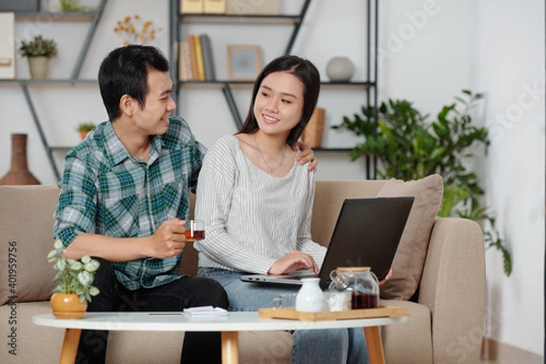 Obraz Smiling Asian man hugging girlfriend working on laptop at home and offering her a cup of tea