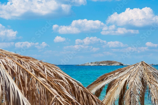 Fototapeta Umbrella made of palm leafs on Tigania beach in Sithonia, Greece with island on a background, sea and sky with clouds