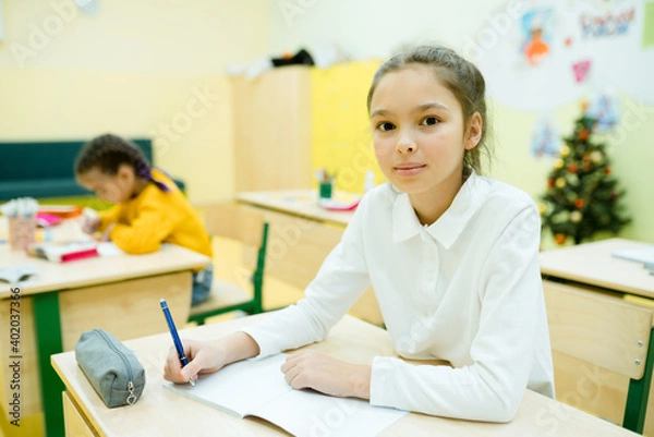 Fototapeta Little girl in a white shirt with gathered hair sits at a desk and writes in a notebook