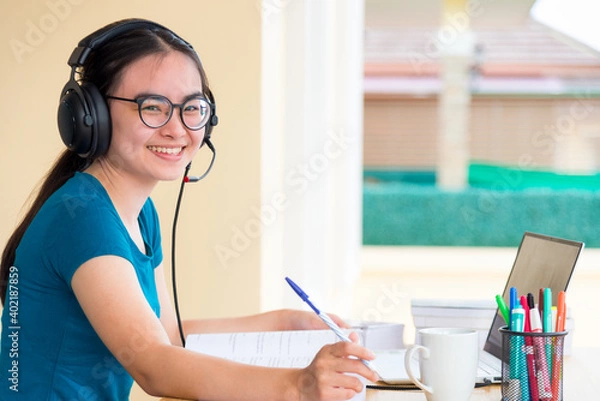 Fototapeta Young Asian teenage girl student with headset glasses is looking while  using a laptop computer study online from school distance education learning from a university class at home for background