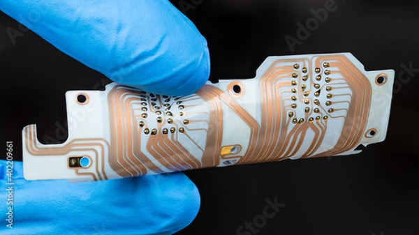 Fototapeta Flex printed circuit board with copper layer in man fingers isolated on black background. Detail of hand in blue glove holding white PCB sheet with integrated connector for computer printer printhead.
