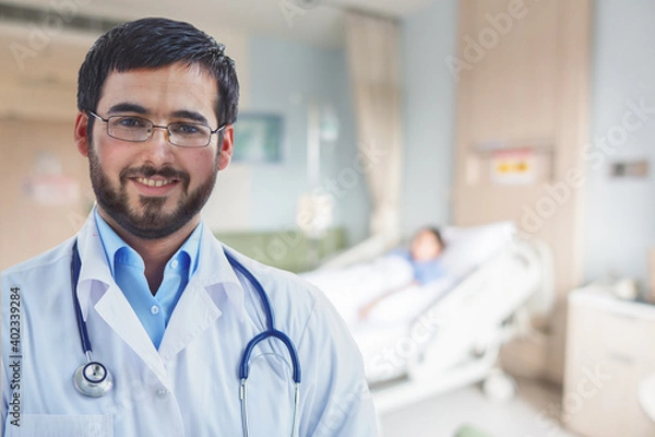 Fototapeta Portrait of smiling Arabian doctor looking at camera with a blurred background of a patient on the bed.
