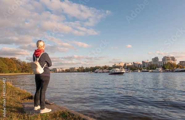 Fototapeta A young woman in casual comfortable clothes and with a backpack stands on the river bank admiring the view.