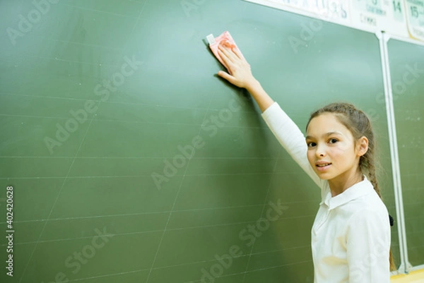 Fototapeta A girl at school stands near the blackboard and uses a rag to wipe it off. School weekdays