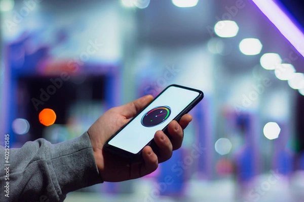Fototapeta A man holds a smartphone with a digital fuel meter on the screen against the background of a night gas station for a car.