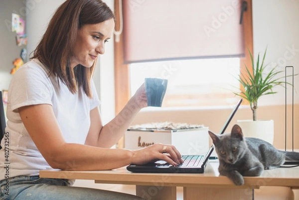 Fototapeta Young woman in white t-shirt sitting with a cat on her lap at the wooden table at home with laptop and notebook, working